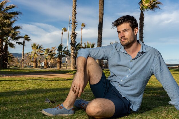 Varón joven hispano vistiendo una camisa azul sentado en un parque con un montón de palmeras en segundo plano.