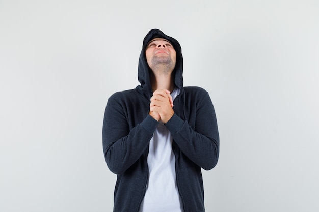 Varón joven juntando las manos en gesto de oración en chaqueta, camiseta y mirando esperanzado, vista frontal.