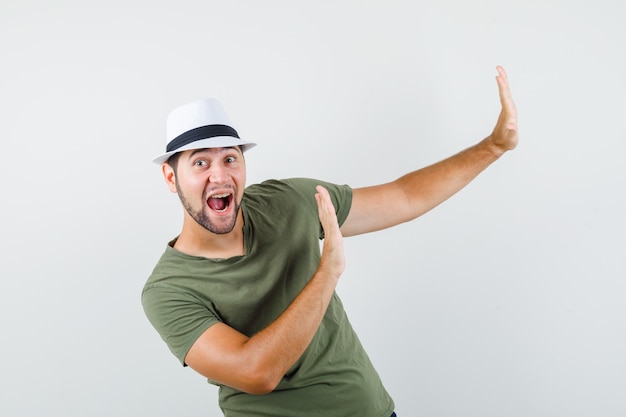 Varón joven levantando las manos de manera preventiva en camiseta verde y sombrero y mirando positivo