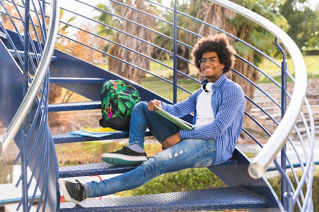 Varón joven con el libro y los bolsos que se relajan en la escalera azul en el parque