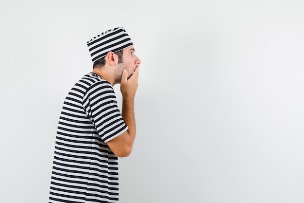 Varón joven con la mano en la boca en camiseta, sombrero y mirando sorprendido. .