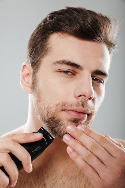 Varón joven masculino mirando a la cámara desvestirse y aislarse en casa con cuidado de la piel mientras se afeita la cara con la recortadora contra la pared gris