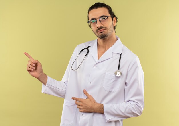 Varón joven médico con gafas ópticas vistiendo túnica blanca con estetoscopio apunta al lado