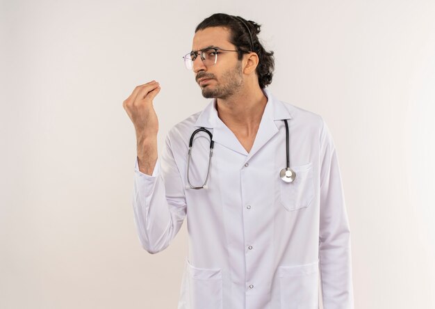 Varón joven médico con gafas ópticas vistiendo túnica blanca con estetoscopio mostrando el gesto de la punta