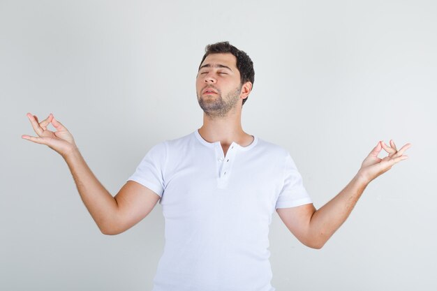 Varón joven meditando con los ojos cerrados en camiseta blanca y mirando tranquilo.