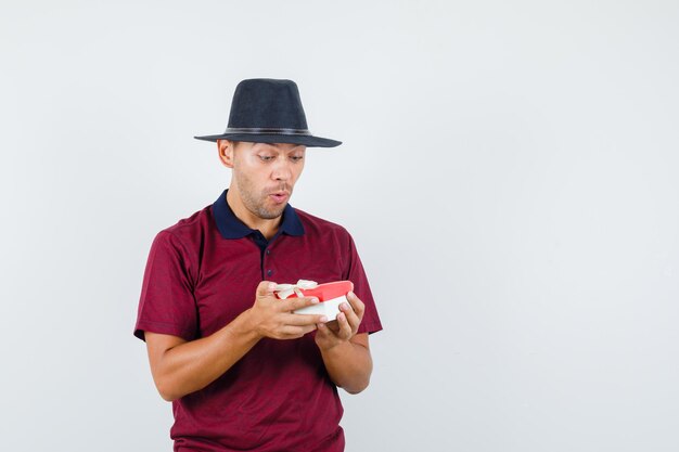 Varón joven mirando regalo en camisa roja y mirando sorprendido, vista frontal. espacio para texto