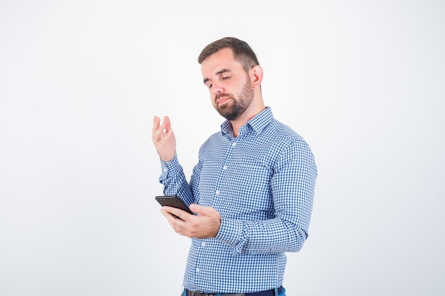 Varón joven mirando el teléfono móvil en camisa, jeans y mirando disgustado, vista frontal.