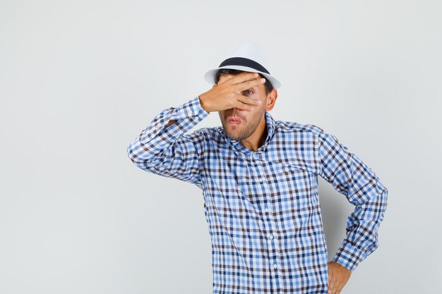 Varón joven mirando a través de los dedos en camisa a cuadros, sombrero y mirando curioso.