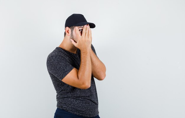 Varón joven mirando a través de los dedos en camiseta y gorra, jeans y mirando curioso