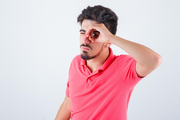 Varón joven mirando a través de los dedos en camiseta y mirando divertido, vista frontal.