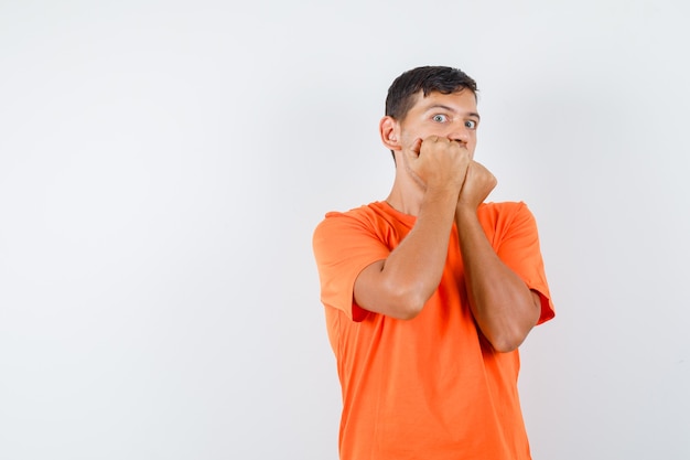 Varón joven mordiendo los puños emocionalmente en camiseta naranja y mirando asustado