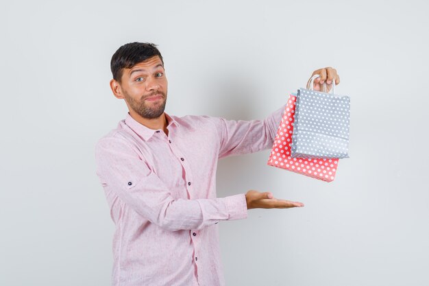 Varón joven mostrando bolsas de papel en camisa y mirando alegre. vista frontal.