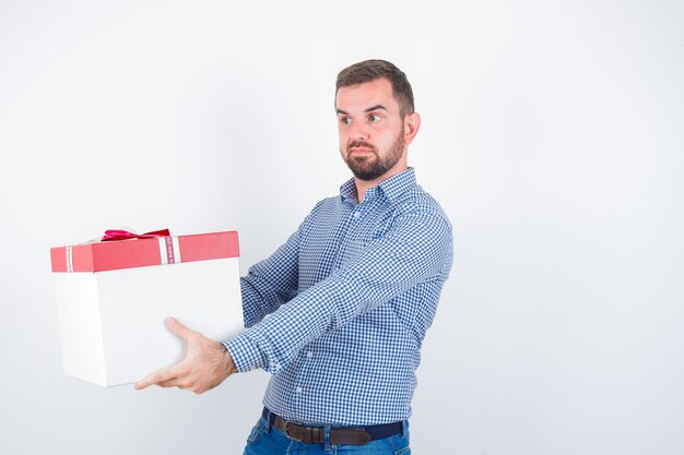 Varón joven mostrando dando gesto mientras sostiene la caja de regalo en camisa, jeans y mirando confiado, vista frontal.