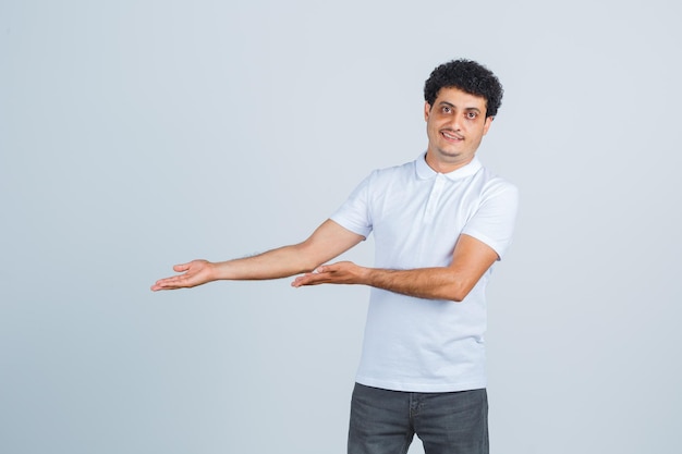 Varón joven mostrando gesto de bienvenida en camiseta blanca, pantalón y mirando confiado. vista frontal.