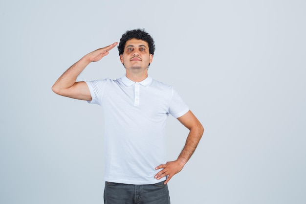 Varón joven mostrando gesto de saludo en camiseta blanca, pantalones y mirando confiado, vista frontal.