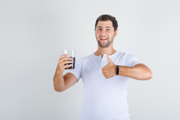 Varón joven mostrando el pulgar hacia arriba con bebida de cola en camiseta blanca y mirando feliz