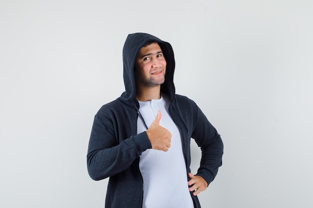 Varón joven mostrando el pulgar hacia arriba en camiseta, chaqueta y mirando confiado, vista frontal.