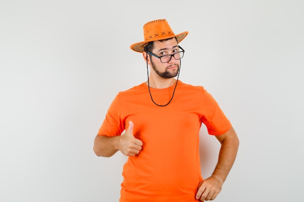 Varón joven mostrando el pulgar hacia arriba en camiseta naranja, sombrero y mirando confiado, vista frontal.