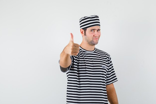 Varón joven mostrando el pulgar hacia arriba en camiseta, sombrero y mirando confiado, vista frontal.
