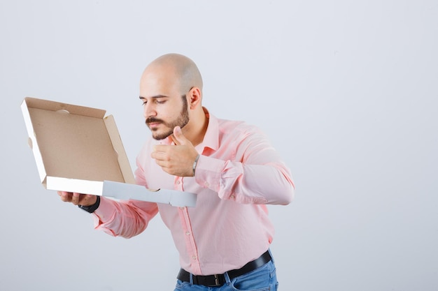 Varón joven oliendo caja de pizza abierta en camisa, jeans y mirando encantador, vista frontal.