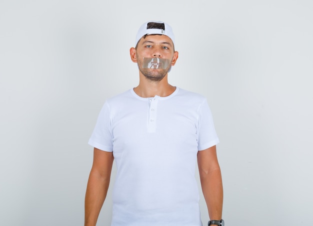 Foto gratuita varón joven de pie con la boca encintada en camiseta blanca, gorra y mirando tranquilo, vista frontal.