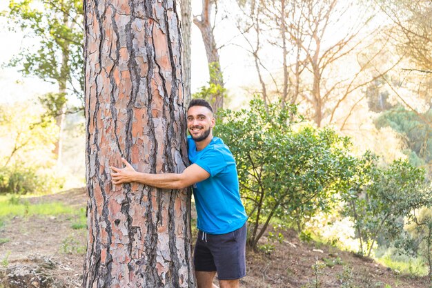 Varón joven de pie en el bosque y abrazando el árbol