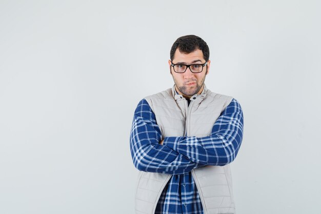 Foto gratuita varón joven de pie con los brazos cruzados en camisa, chaqueta, gafas y mirando con cuidado. vista frontal.