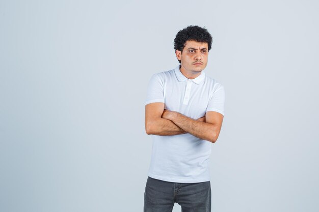 Varón joven de pie con los brazos cruzados en camiseta blanca, pantalones y mirando pensativo, vista frontal.