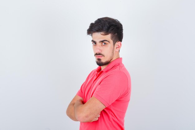 Varón joven de pie con los brazos cruzados en camiseta rosa y mirando confiado, vista frontal.