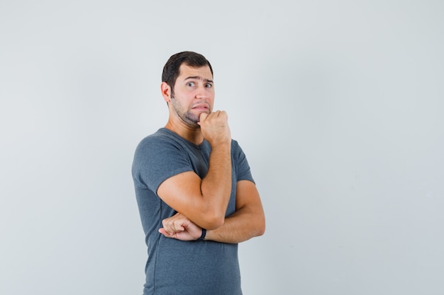 Varón joven de pie en pose de pensamiento en camiseta gris y mirando ansioso