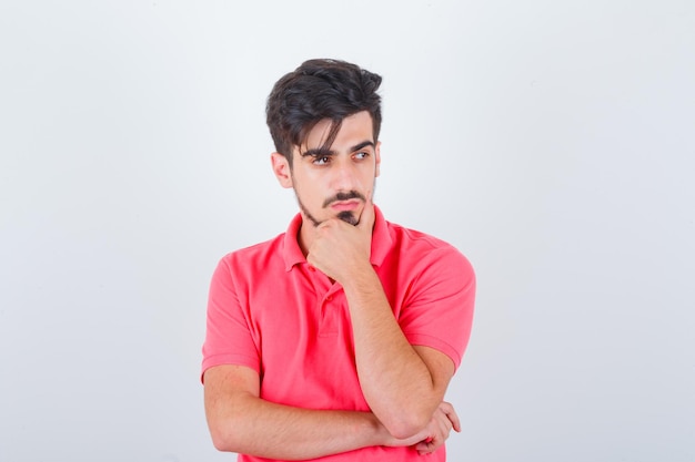 Varón joven de pie en pose de pensamiento en camiseta y mirando sensible, vista frontal.