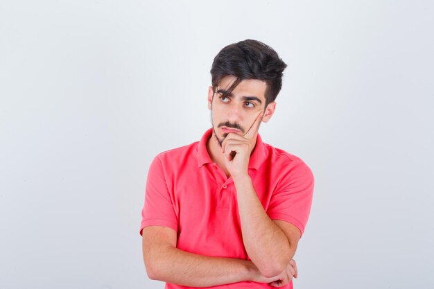 Varón joven de pie en pose de pensamiento en camiseta y mirando sensible, vista frontal.