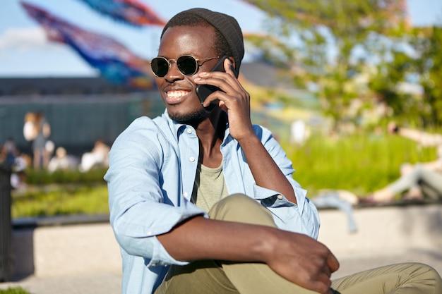 Varón joven positivo con piel oscura, sonriendo ampliamente mientras conversa con su mejor amigo, hablando por teléfono inteligente mientras descansa al aire libre. Personas, comunicación, concepto de tecnología.