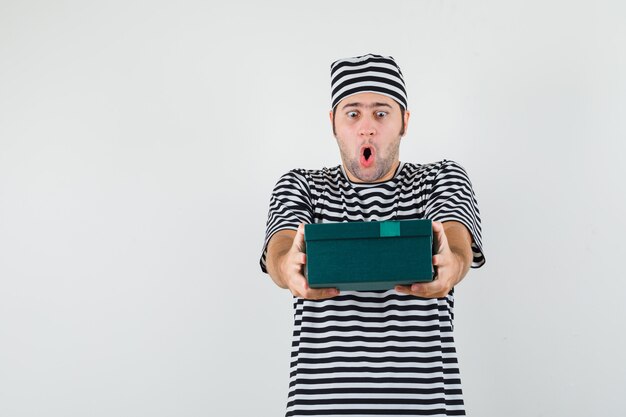 Varón joven presentando caja de regalo en camiseta, sombrero y mirando sorprendido, vista frontal.