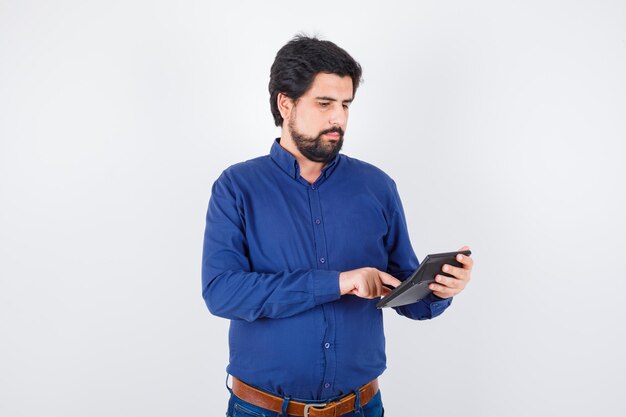 Varón joven presionando los botones de la calculadora en camisa azul real y mirando con cuidado, vista frontal.