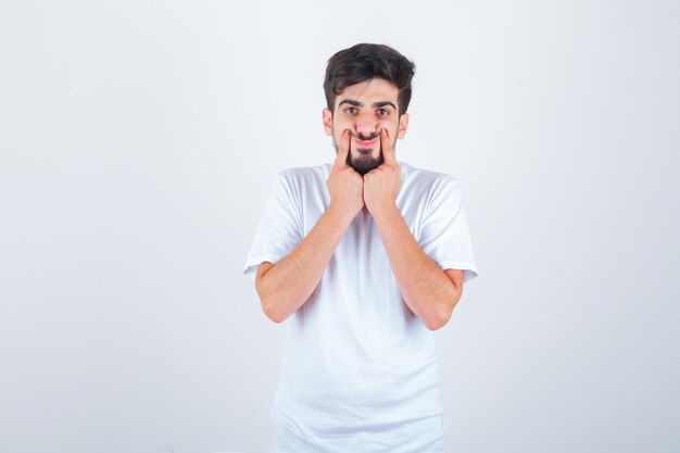 Varón joven presionando los dedos en las mejillas en camiseta y luciendo lindo. vista frontal.