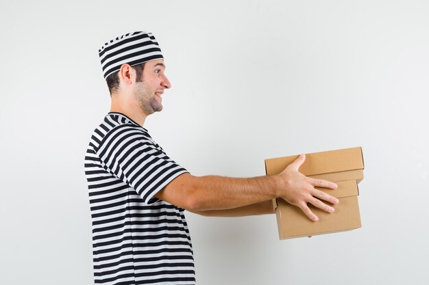 Varón joven que entrega cajas de cartón en camiseta, sombrero y mirando alegre. .