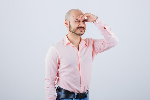 Varón joven que lleva a cabo la mano en la frente en camisa, pantalones vaqueros y que parece alegre. vista frontal.