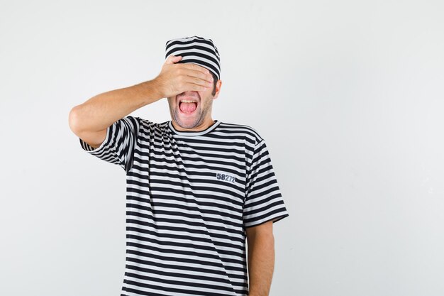 Varón joven que lleva a cabo la mano en los ojos en camiseta, sombrero y que parece feliz. vista frontal.