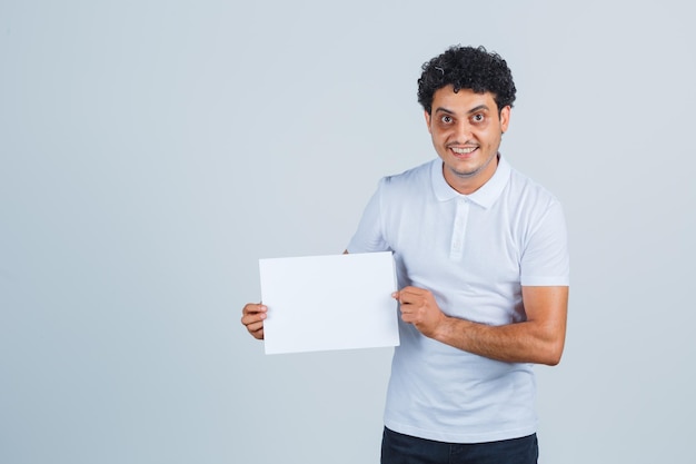 Varón joven que mantiene una hoja de papel en blanco con una camiseta blanca, pantalones y una mirada segura, vista frontal.