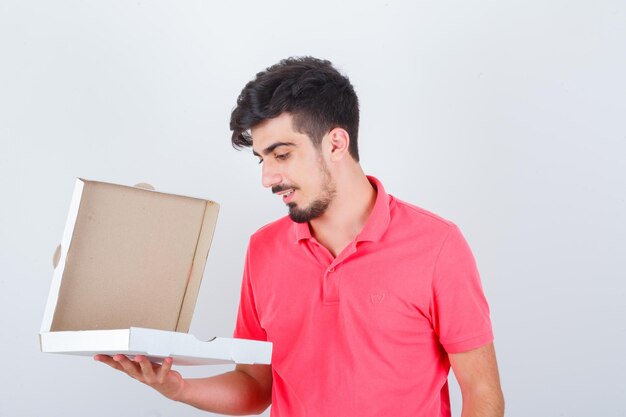 Varón joven que mira la caja abierta de la pizza en la camiseta y que mira vacilante. vista frontal.