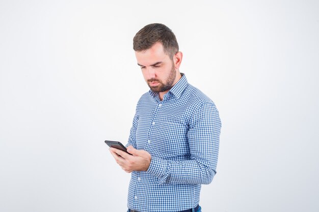 Varón joven que mira el teléfono móvil en camisa, pantalones vaqueros y que parece serio. vista frontal.