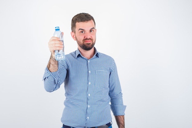 Varón joven que muestra una botella de agua de plástico en camisa, jeans y parece seguro. vista frontal.