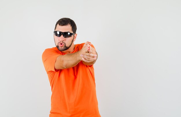Varón joven que muestra el gesto del arma apuntado en la camiseta naranja y que mira confiado, vista frontal.