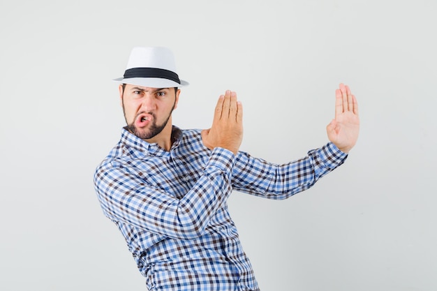 Foto gratuita varón joven que muestra gesto de chuleta de karate en camisa a cuadros, sombrero y con enojo. vista frontal.