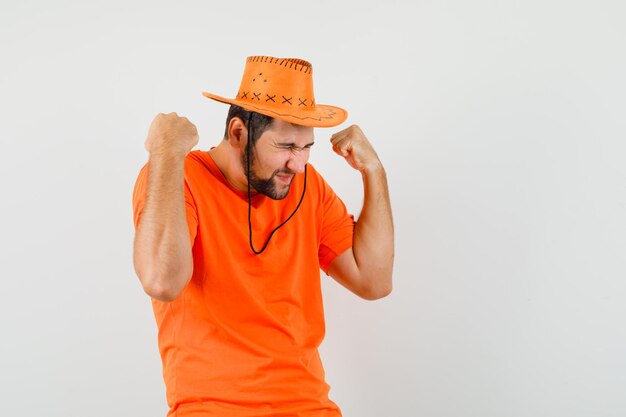 Varón joven que muestra gesto de éxito en camiseta naranja, sombrero y mirando feliz, vista frontal.