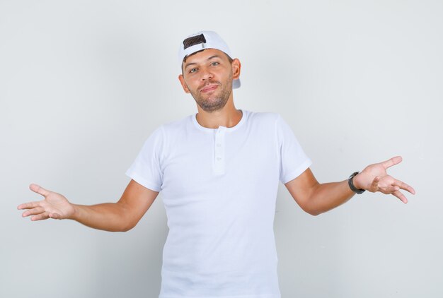 Varón joven que muestra un gesto de impotencia con las manos en la camiseta blanca, gorra y mirando confundido, vista frontal.