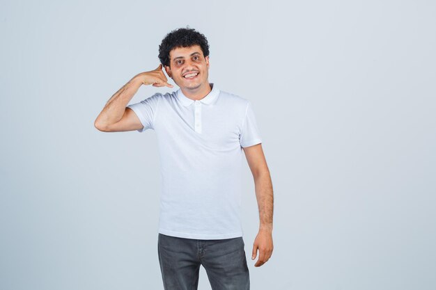 Varón joven que muestra el gesto del teléfono en camiseta blanca, pantalones y mirando alegre, vista frontal.