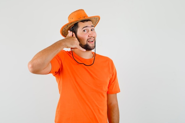 Varón joven que muestra el gesto del teléfono en camiseta naranja, sombrero y mirando alegre, vista frontal.