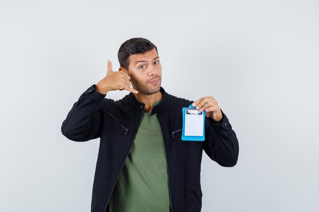 Varón joven que muestra el gesto del teléfono mientras sostiene el portapapeles en la camiseta, la chaqueta y parece útil, vista frontal.